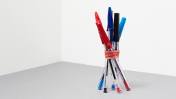 6 office pens of different colors, rubber banded together, and upright.